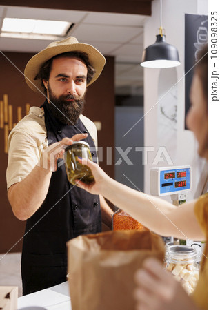 Woman at checkout counter in zero waste shop, buying locally sourced ecofriendly vegetables from happy storekeeper. Client purchasing pickles jar in local neighborhood store using renewable energy 109098325