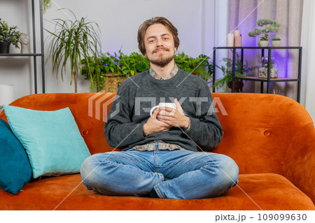Attractive smiling Caucasian young man drinking a cup of coffee or herbal tea sitting at home sofa Attractive smiling Caucasian young man drinking a cup of coffee or herbal tea sitting at home sofa 109099630