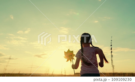 Little girl runs to horizon holding paper windmill across meadow at sunset light Little girl runs to horizon holding paper windmill across meadow at sunset light 109109230