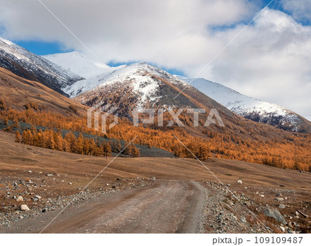 Long and winding road. Picturesque autumn landscape with a rocky dirt road through the steppe in the mountains, golden autumn trees on the slopes and the first snow on top mountains. Long and winding road. Picturesque autumn landscape with a rocky dirt road through the steppe in the mountains, golden autumn trees on the slopes and the first snow on top mountains. 109109487
