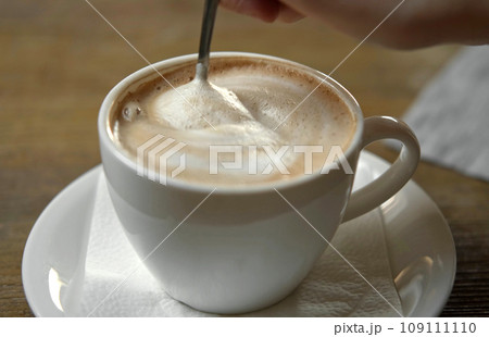 Hand is Mixing the Milk Foam of Latte. Person Stirring Cappuccino with Steel Spoon. Hot Coffee in a White Classic Ceramic Cup on Wooden Table. Modern Coffee shop, Cafeteria. Thick Froth. Close-up View 109111110