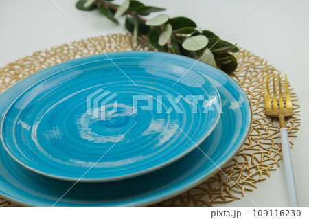 Bright blue serving plates stand on golden napkins, a fork of gold and white color is located on the side, a plant is visible behind the plate, a side view on a white background. The concept of 109116230