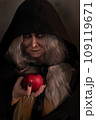 Stepmother witch gives poisoned red apple.Woman as witch in black offers red apple as symbol of temptation, poison. Fairy tale, white snow wizard concept. Spooky Halloween, cosplay. 109119671