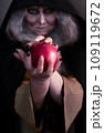 Stepmother witch gives poisoned red apple.Woman as witch in black offers red apple as symbol of temptation, poison. Fairy tale, white snow wizard concept. Spooky Halloween, cosplay. 109119672