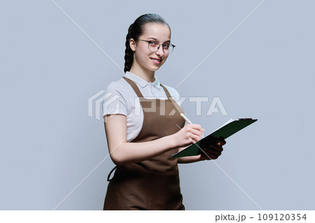 Young female worker in apron writing on clipboard paper, on gray background Young female worker in apron writing on clipboard paper, on gray background 109120354
