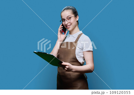 Young female worker in apron taking order by phone, writing, on blue background Young female worker in apron taking order by phone, writing, on blue background 109120355
