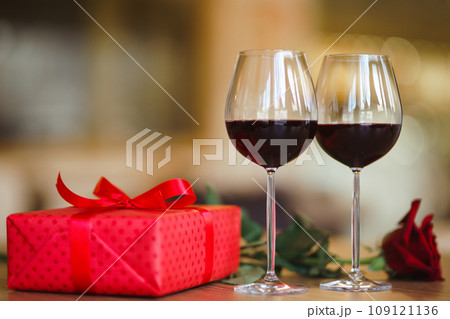 Valentine's day celebration concept. Two glasses of wine, red rose red gift box on wooden table. Valentine's day celebration concept. Two glasses of wine, red rose red gift box on wooden table. 109121136