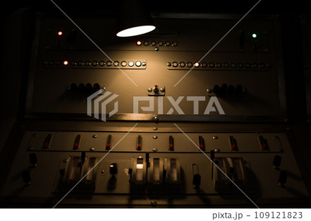 Antique control panel of a nuclear power plant is illuminated in the dark by a lamp. Vintage control panel with buttons Antique control panel of a nuclear power plant is illuminated in the dark by a lamp. Vintage control panel with buttons 109121823