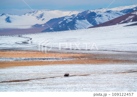 Beautiful Mongolian winter landscape snow mountain with a small figure of a riders on a black horses galloping through a wide snow-covered valley against the backdrop of high mountains. Beautiful Mongolian winter landscape snow mountain with a small figure of a riders on a black horses galloping through a wide snow-covered valley against the backdrop of high mountains. 109122457