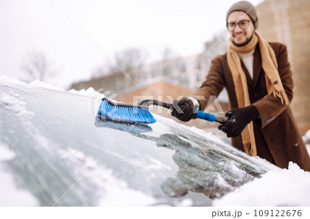 Stylish man in a coat and scarf clears snow from his car during winter snowfall. Seasonality concept 109122676