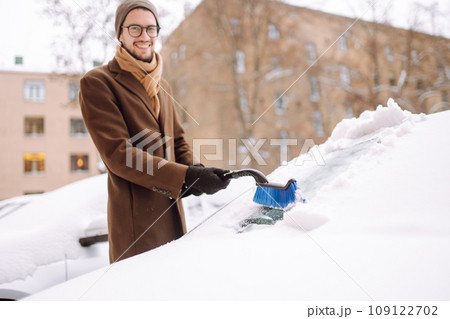 Stylish man in a coat and scarf clears snow from his car during winter snowfall. Seasonality concept 109122702