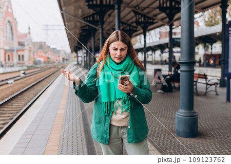 Angry redhead girl with smart phone standing at railroad station platform. Negative people emotion. Travel to vacation by train. Young redhead woman waiting train with backpack and using smart phone. 109123762