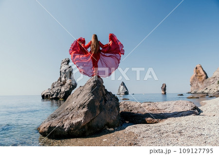 Woman travel sea. Young Happy woman in a long red dress posing on a beach near the sea on background of volcanic rocks, like in Iceland, sharing travel adventure journey 109127795
