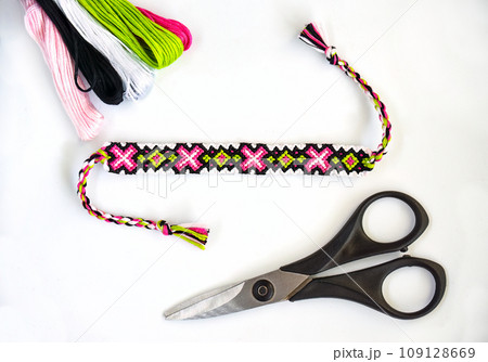 Woven handmade DIY friendship bracelets with bright pink and green pattern next to skeins of thread and scissors on white background 109128669