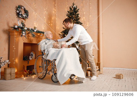 Bearded man and his father celebrating Christmas together. Man and his old father on a wheelchair sitting near Christmas tree. Son giving a father gift box. 109129606