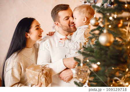 Young family celebrating Christmas together. Brunette mother, bearded father and blonde son standing near Christmas tree and holding a gift box. Family wearing beige knitted clothes. 109129718