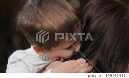 Mom stands with son in arms and covers with kisses in cheek. Mom amidst park setting holds son filling air with warmth. Mom in ambiance of park with son in arms shares moment of creating bond Mom stands with son in arms and covers with kisses in cheek. Mom amidst park setting holds son filling air with warmth. Mom in ambiance of park with son in arms shares moment of creating bond 109132411