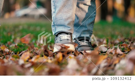 Legs of young boy sauntering along lawn. Legs of boy doing steps on park lawn. Legs of young boy walking on way across lawn with vivid thick carpet of leaves creating pleasant and playful scene 109132412