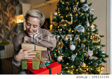 Portrait of happy elegant middle-aged woman holding big pile of wrapped gifts, sitting by decorated Christmas tree, smiling looking with happiness expression. Concept of home festive atmosphere. 109133140