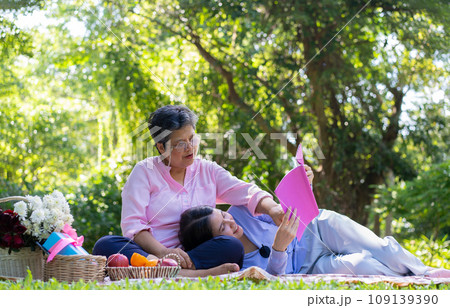 Asian daughter Sleeping on mother lap and reading book on picnic mat in the park. A happy senior woman talks with her daughter. Concept of healthcare and elderly caregiver support service. 109139390