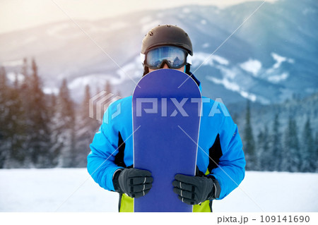 Portrait Of Man On The Background Of Mountains And Blue Sky, Holding Snowboard And Wearing Ski Glasses. Ski Goggles Of Snowboarder With The Reflection Of Snowed Mountains. Winter Sports Portrait Of Man On The Background Of Mountains And Blue Sky, Holding Snowboard And Wearing Ski Glasses. Ski Goggles Of Snowboarder With The Reflection Of Snowed Mountains. Winter Sports 109141690