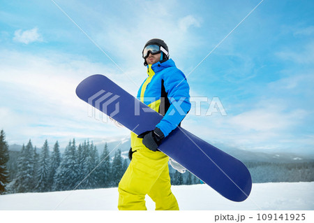 Portrait Of Man On The Background Of Mountains And Blue Sky, Holding Snowboard And Wearing Ski Glasses. Ski Goggles Of Snowboarder With The Reflection Of Snowed Mountains. Winter Sports 109141925