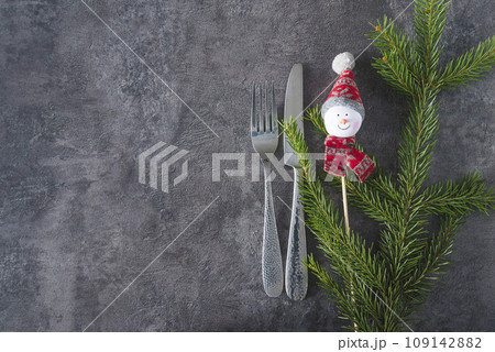 Christmas holiday place setting; Fork and knife, fir twig and snowman decoration on the gray textured background; copy space, flat lay Christmas holiday place setting; Fork and knife, fir twig and snowman decoration on the gray textured background; copy space, flat lay 109142882