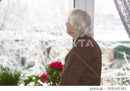Old lonely woman standing near the window in his house and looking and dreaming at winter 109145361