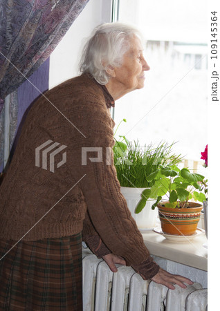 Old lonely woman standing near the window in his house and looking and dreaming at winter 109145364