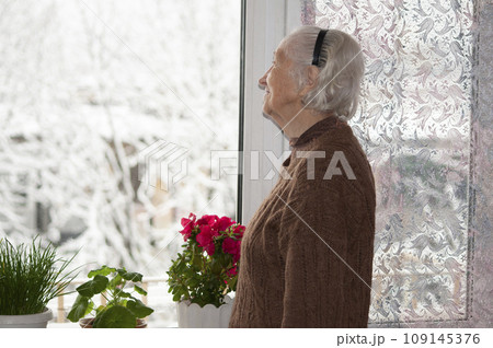 Old lonely woman standing near the window in his house and looking and dreaming at winter 109145376