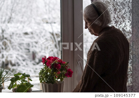 Old lonely woman standing near the window in his house and looking and dreaming at winter 109145377