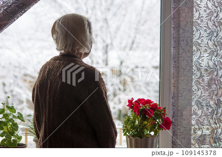 Old lonely woman standing near the window in his house and looking and dreaming at winter 109145378