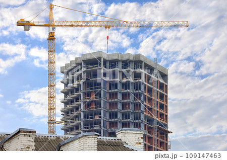A tower crane is erecting a new residential building using the monolithic frame method against the background of the old roof of an outdated house. A tower crane is erecting a new residential building using the monolithic frame method against the background of the old roof of an outdated house. 109147463