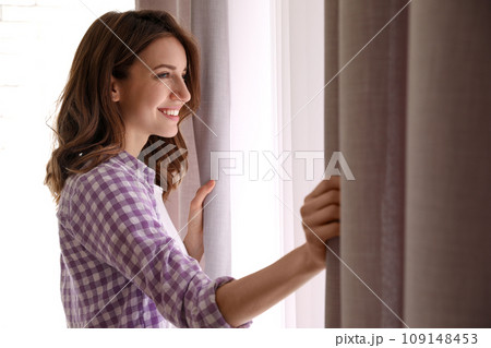 Woman opening window curtains at home in morning 109148453