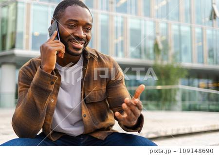 African american man talking on smartphone on street in city outdoor. Man with cell phone chatting with friends. Smiling person making answering call by cellphone. Guy having conversation by mobile African american man talking on smartphone on street in city outdoor. Man with cell phone chatting with friends. Smiling person making answering call by cellphone. Guy having conversation by mobile 109148690