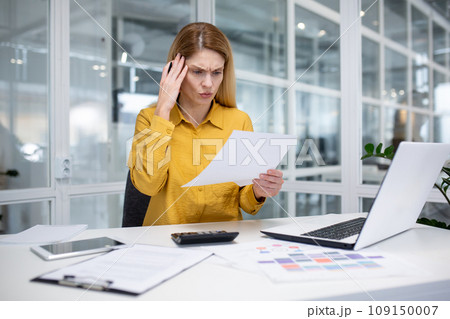Serious young business woman working in the office at the laptop, holding documents in her hands and holding her head worriedly. 109150007
