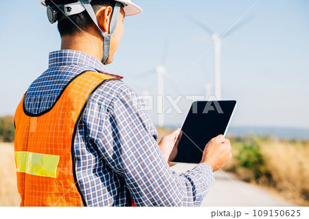An engineer tablet in hand works at windmill farm. Generating clean energy the technician's expertise guarantees turbine efficiency. Global innovation in electricity control a confident stance. 109150625