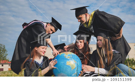 Graduates in black robes examine a geographical globe sitting on the grass. 109151399