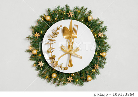 Beautiful Christmas table setting with gold cutlery, gold ornaments and festive wreath on white background. New Year party. Top view, flat lay. Beautiful Christmas table setting with gold cutlery, gold ornaments and festive wreath on white background. New Year party. Top view, flat lay. 109153864