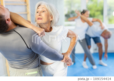 Elderly woman practices strikes and submissions against an attacking man during self-defense lesson in gym 109154414