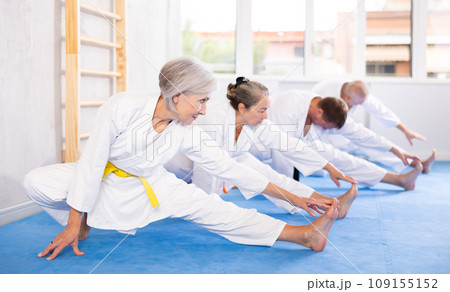 Group of mature women and men in kimonos doing muscle stretching before karate or judo class 109155152