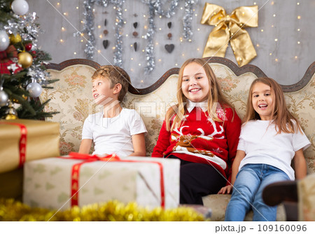 Happy brother with two sisters are sitting on sofa together with gift boxes next to the new year tree 109160096