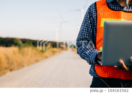 Engineer holding laptop at wind turbines ensuring efficiency. Service technician focuses on turbine performance. Landscape development innovated for windmill service and quality assurance. Engineer holding laptop at wind turbines ensuring efficiency. Service technician focuses on turbine performance. Landscape development innovated for windmill service and quality assurance. 109163562