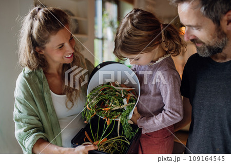 Girl helping parents put kitchen waste, peel and leftover vegetables scraps into kitchen compostable waste. Concept of composting kitchen biodegradable waste. Girl helping parents put kitchen waste, peel and leftover vegetables scraps into kitchen compostable waste. Concept of composting kitchen biodegradable waste. 109164545