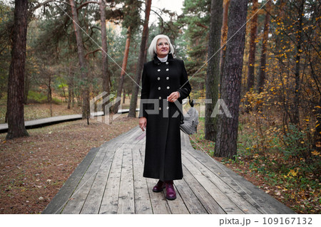 Graceful mature woman dressed in chic black coat, stroll through enchanting forest, reveling in splendor of nature with serene smile, grace and relaxation Graceful mature woman dressed in chic black coat, stroll through enchanting forest, reveling in splendor of nature with serene smile, grace and relaxation 109167132