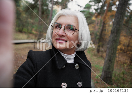 Authentic selfie portrait of senior 60 year old woman with gray hair and glasses against background of nature forest. Real person, natural color Authentic selfie portrait of senior 60 year old woman with gray hair and glasses against background of nature forest. Real person, natural color 109167135