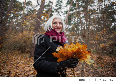 Caucasian happy senior woman with autumn leaves smiling in nature forest. Portrait active mature female with gray hair feels good outdoors. Fall colors, Elderly happiness and Wellbeing Caucasian happy senior woman with autumn leaves smiling in nature forest. Portrait active mature female with gray hair feels good outdoors. Fall colors, Elderly happiness and Wellbeing 109167143