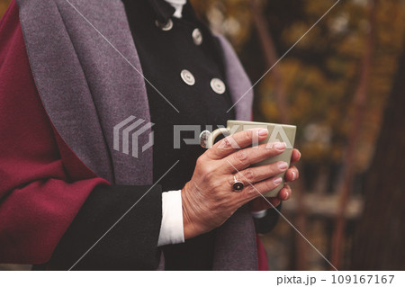 Close up hands of elderly woman, dressed comfortably, holding cup with coffee or tea against backdrop of autumn nature. Relaxing atmosphere, warm colors Close up hands of elderly woman, dressed comfortably, holding cup with coffee or tea against backdrop of autumn nature. Relaxing atmosphere, warm colors 109167167