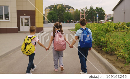 Junior children with schoolbags walk together after school lessons. Happy children with backpacks leave school yard joining hands. Little children go home after schooling in modern city street Junior children with schoolbags walk together after school lessons. Happy children with backpacks leave school yard joining hands. Little children go home after schooling in modern city street 109168246