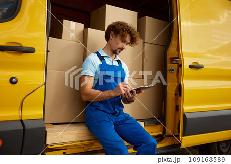 Young courier in overalls sitting at minivan filling in delivery form 109168589